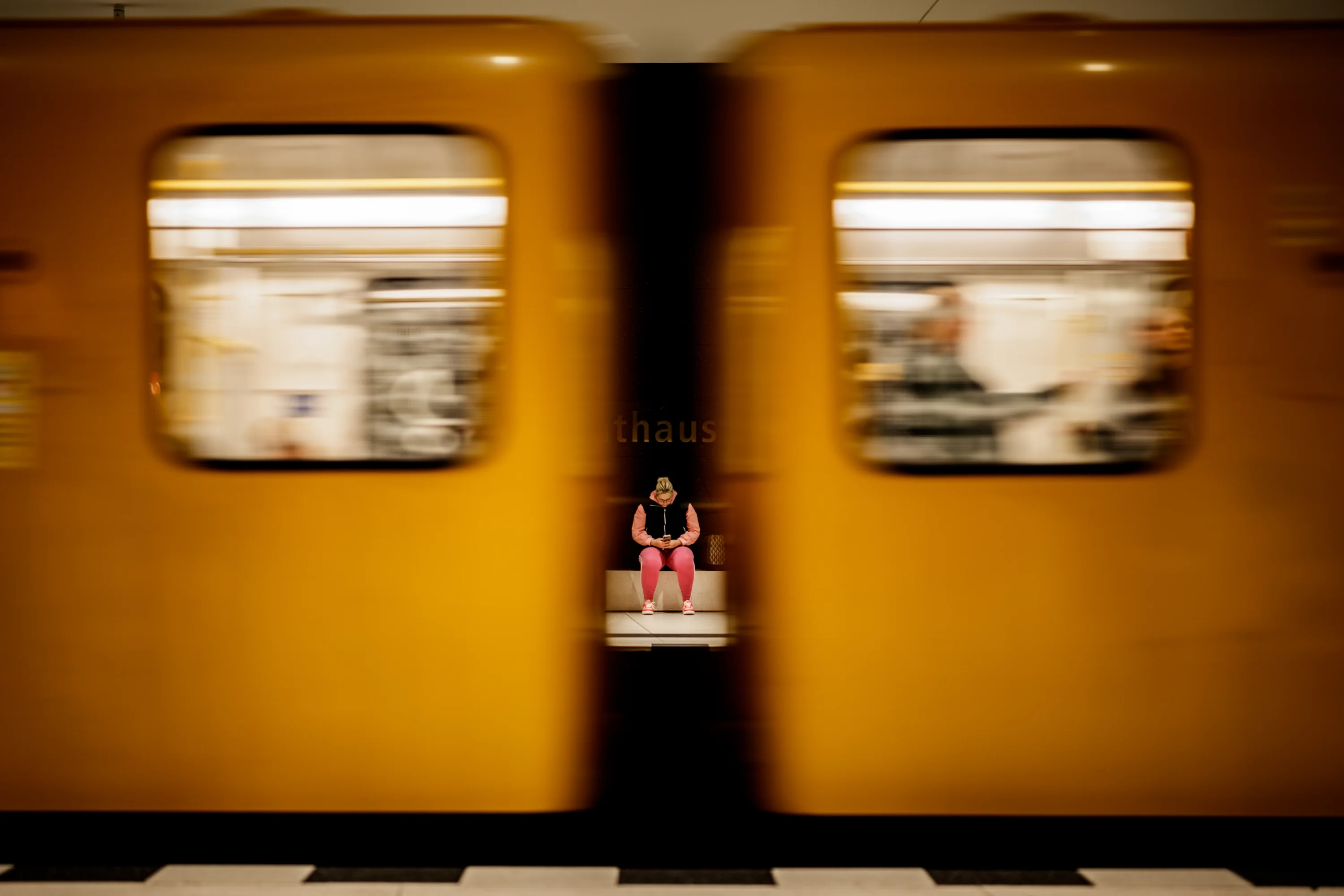 Eine Frau in Pink sitzt still auf einer Bank im Berliner U-Bahnhof Rotes Rathaus, eingerahmt von den verwischten gelben Wagen einer vorbeifahrenden U-Bahn