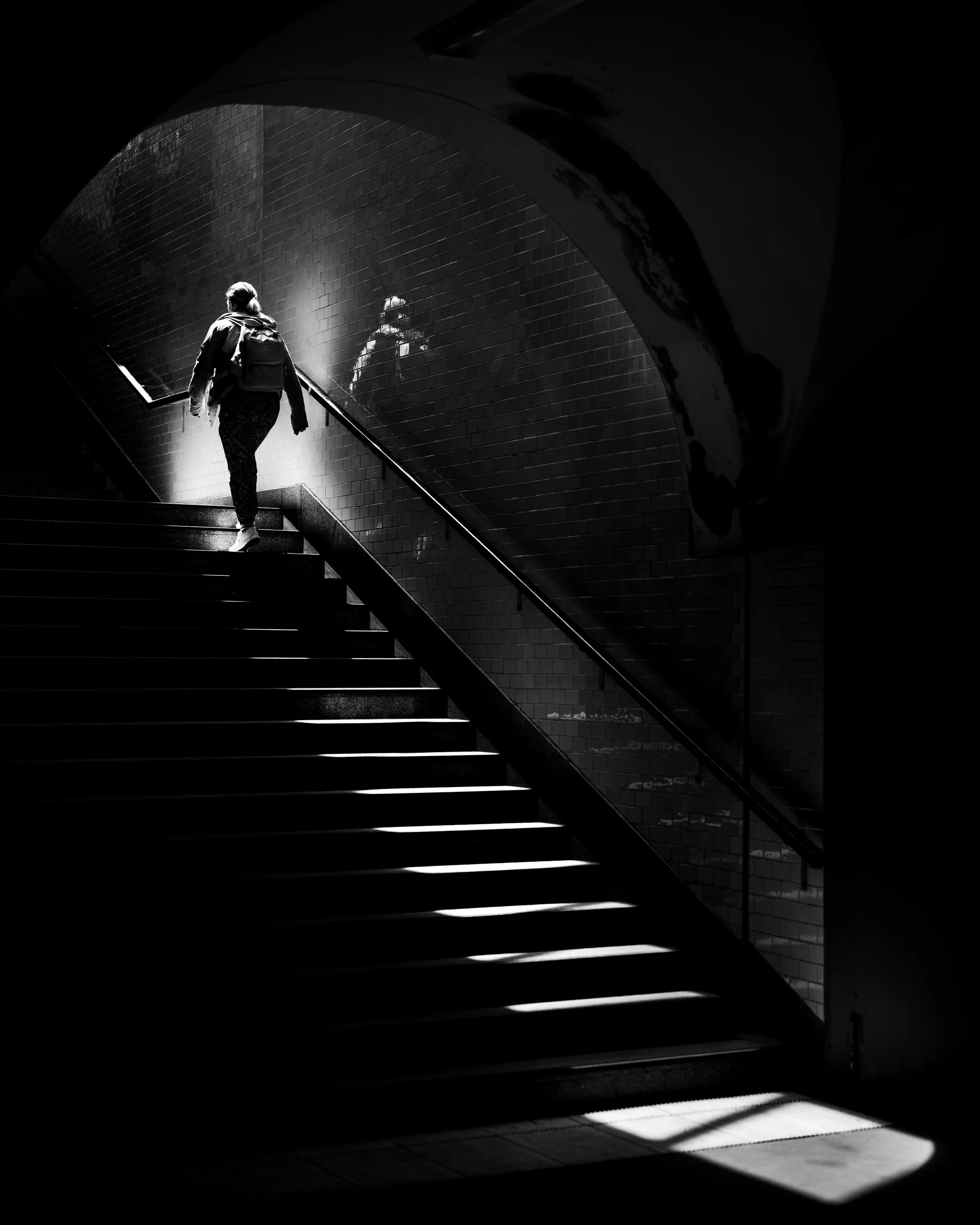 Black-and-white image of a woman climbing the stairs of Gleisdreieck station in Berlin, her faint reflection on the tiled wall beside her