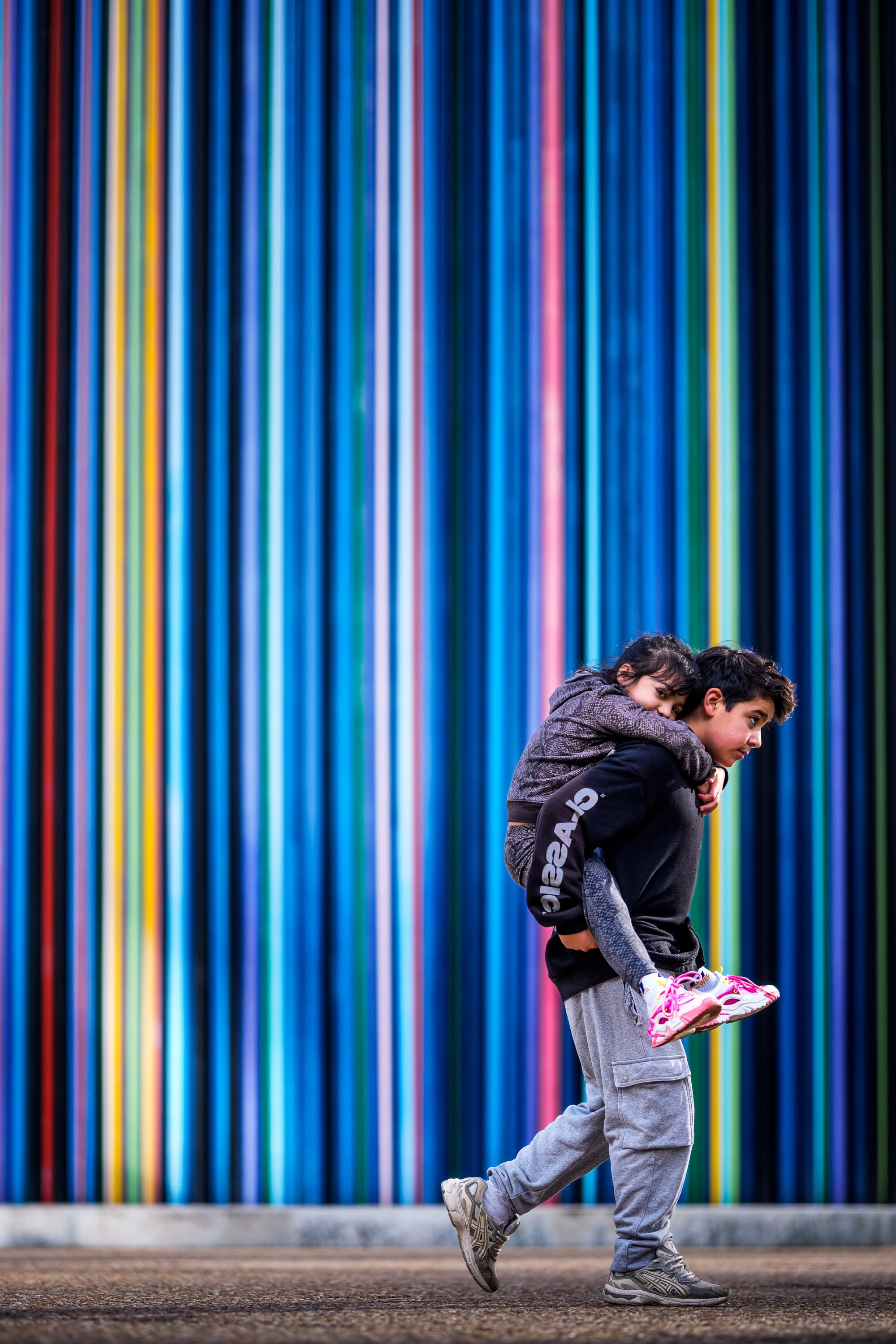 Boy carrying child on his back in front of colorful Moretti Columns at La Défense, Paris — vibrant street photography