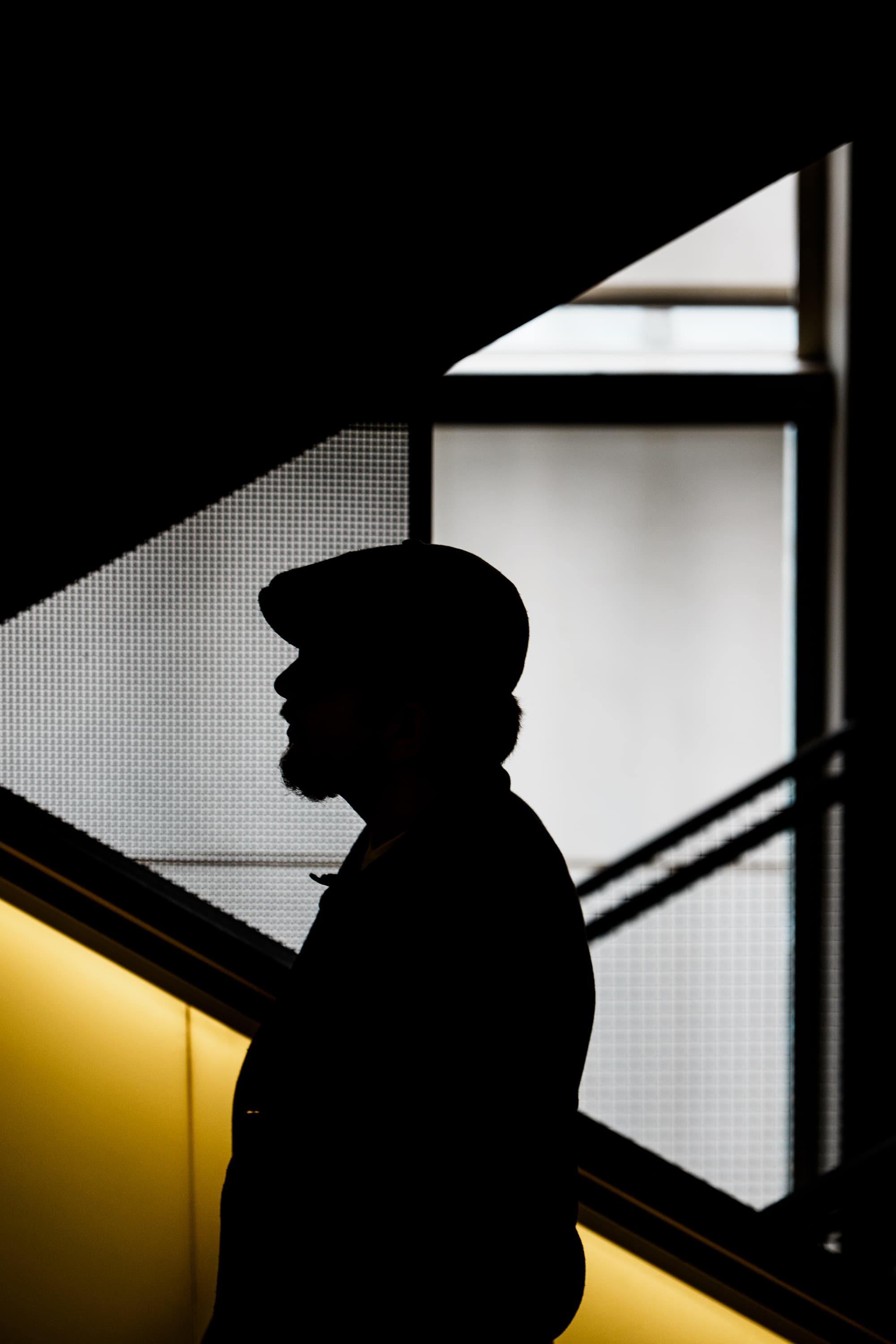Silhouette of a man with cap on escalator in yellow underground light — moody urban street photography