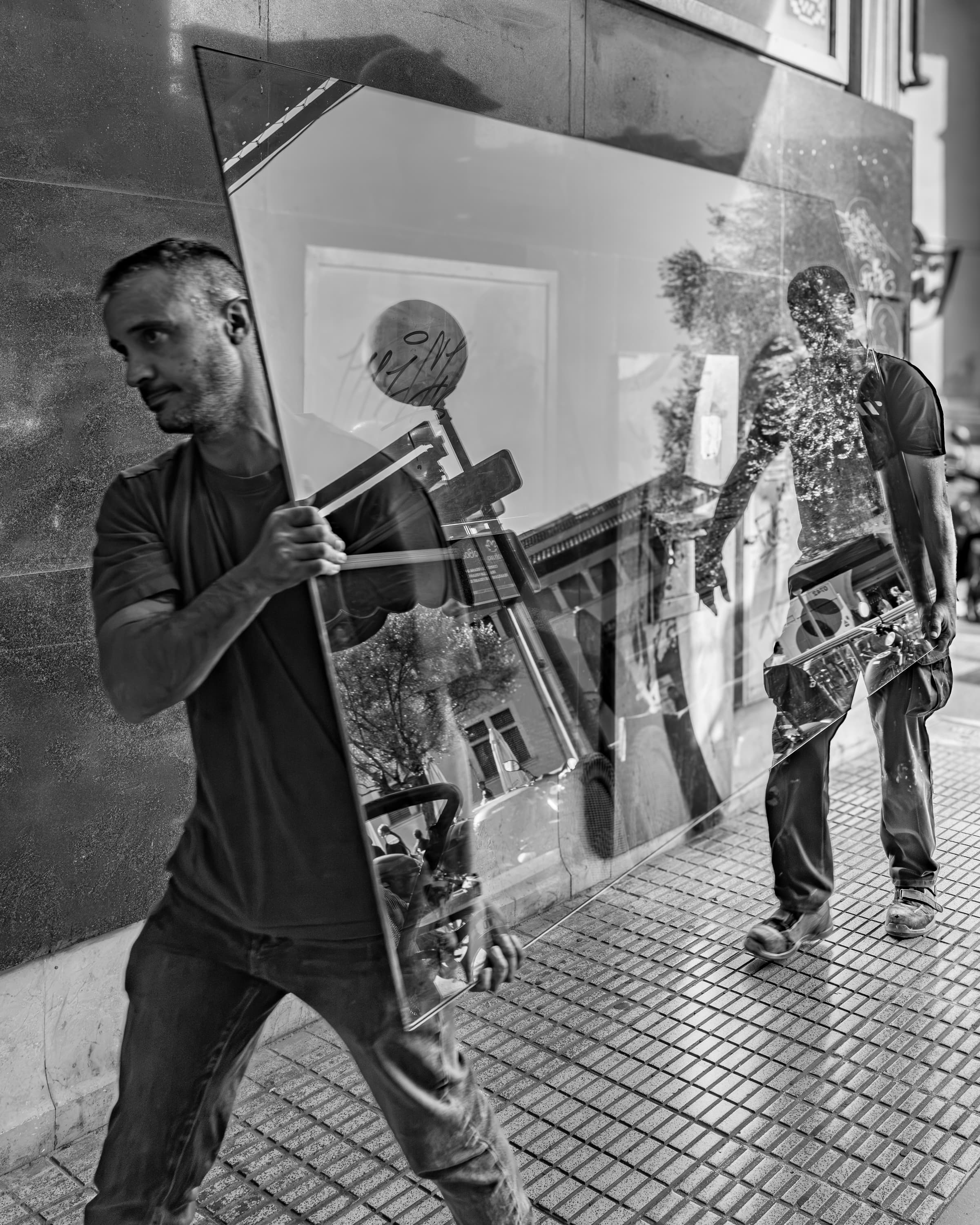 Two workers carrying a large glass pane reflecting the street scene — black and white candid street photography