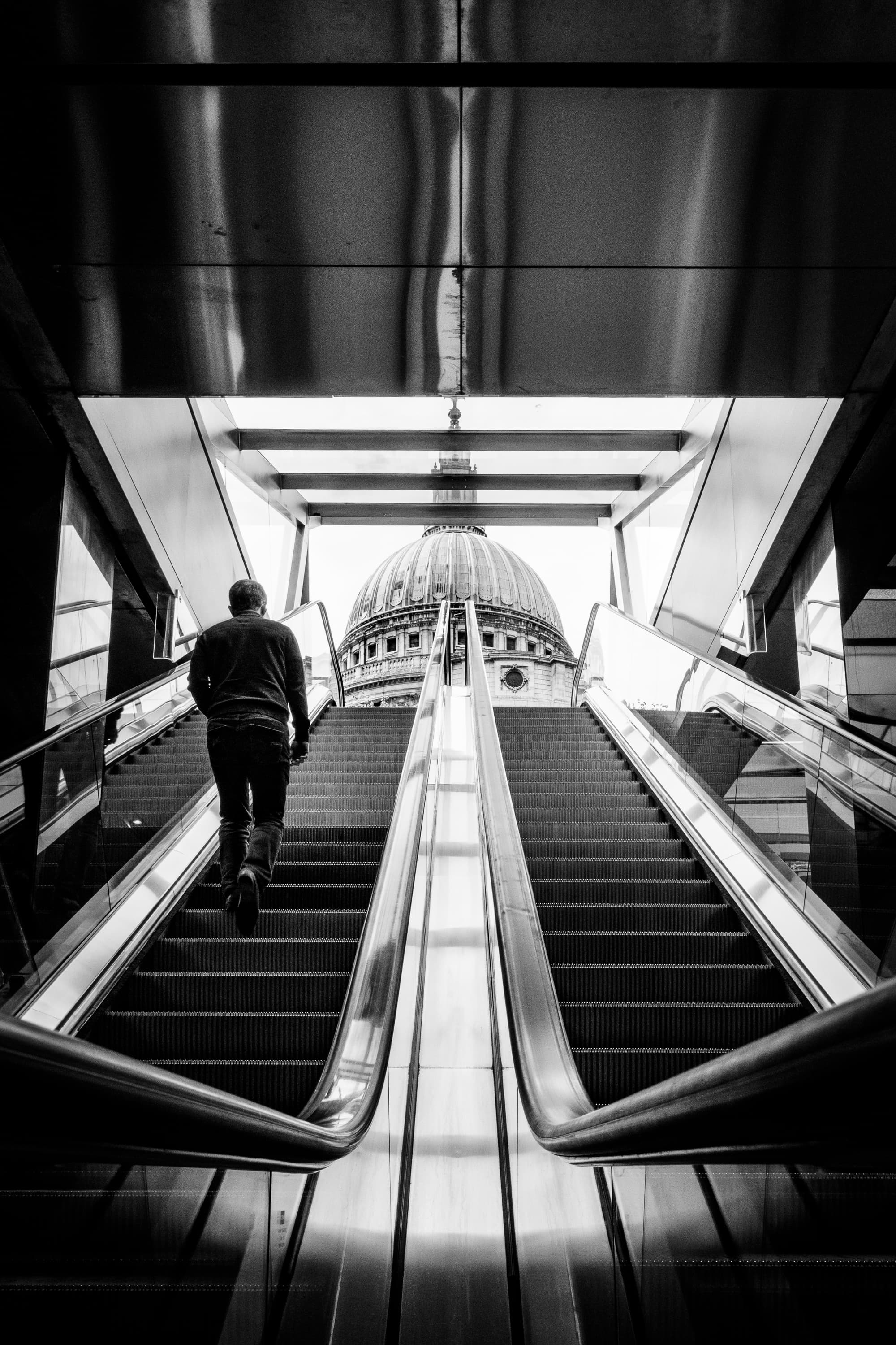 Man ascending escalator towards St Paul's Cathedral dome, London — symmetrical black and white street photography