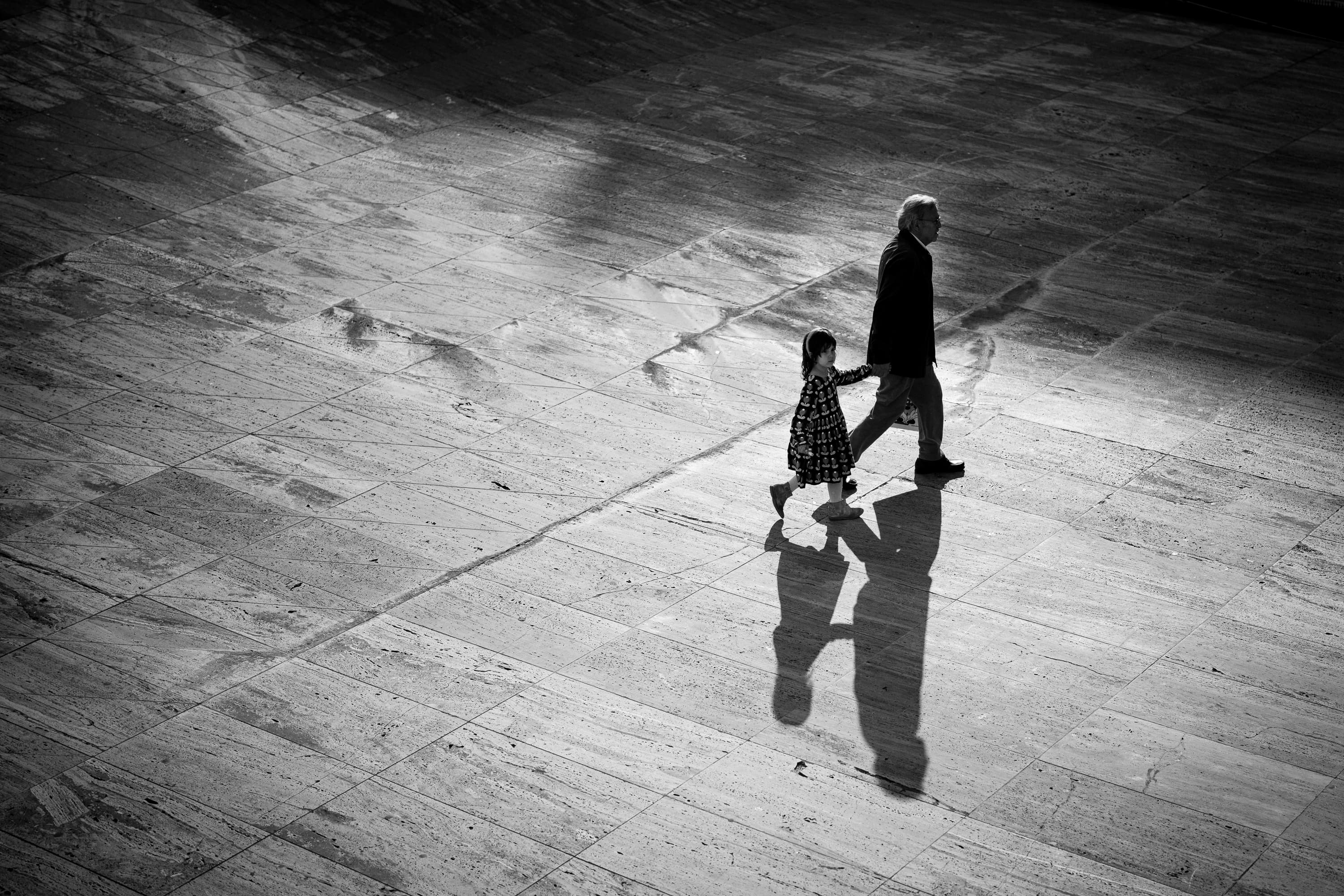 Grandfather and granddaughter walking hand in hand across sunlit plaza, aerial view with long shadows — black and white