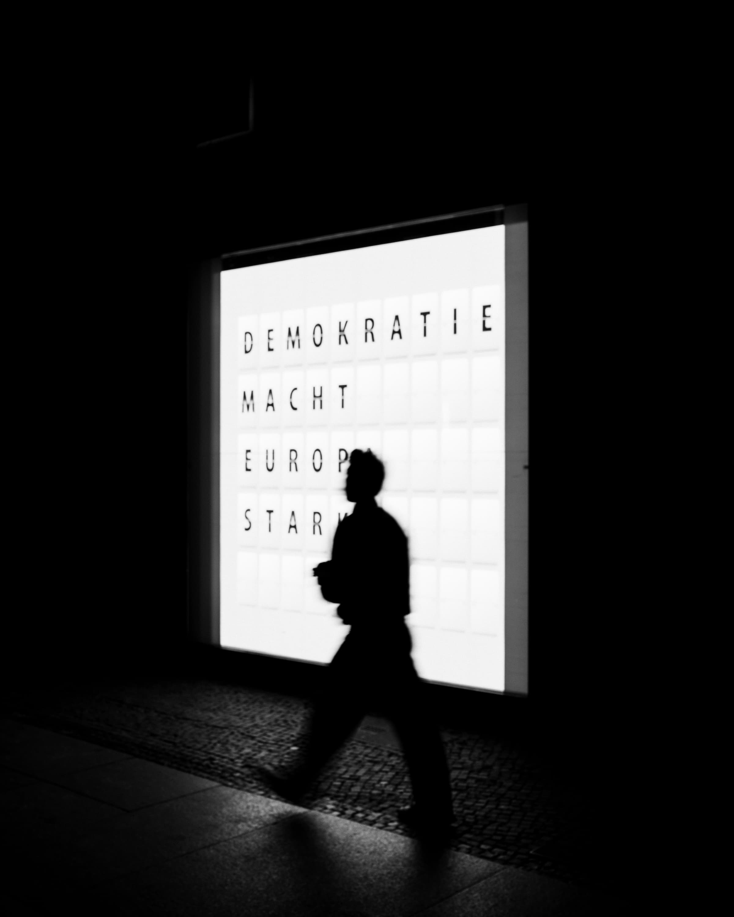 Silhouette walking past illuminated sign reading Demokratie Macht Europa Stark at night, Berlin — black and white