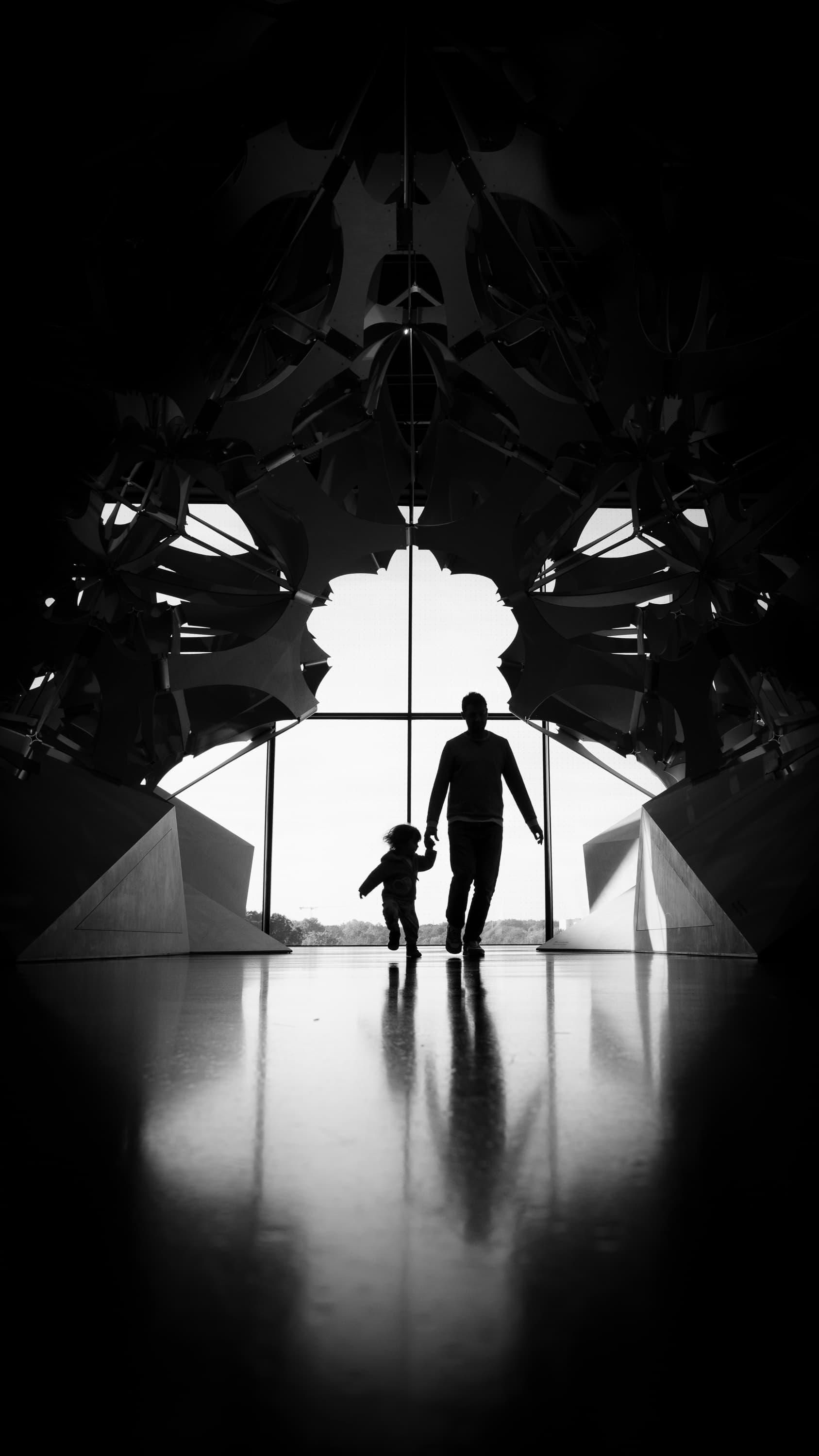 Father and child silhouettes walking into the light through Fondation Louis Vuitton architecture, Paris — black and white