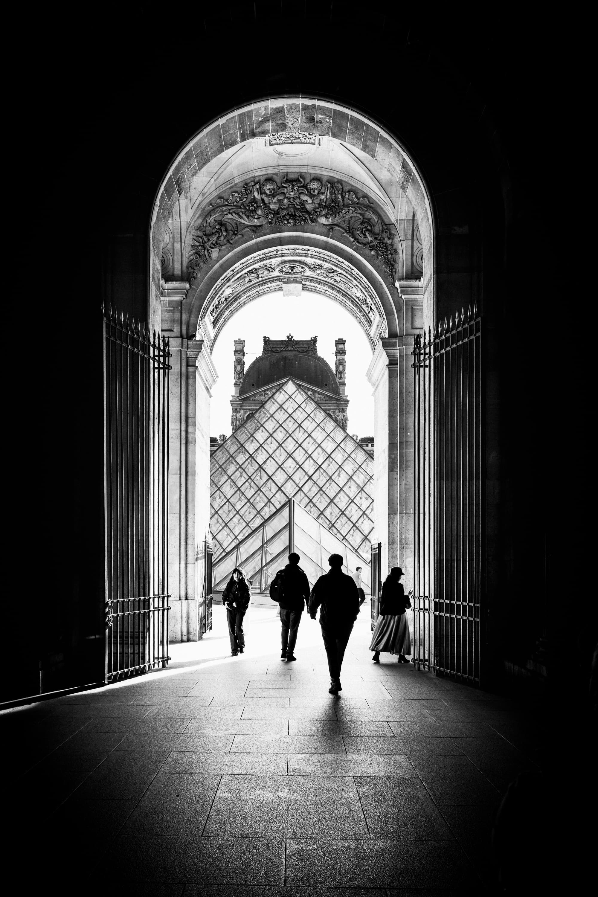 Silhouettes walking through Louvre archway towards the glass pyramid, Paris — black and white street photography