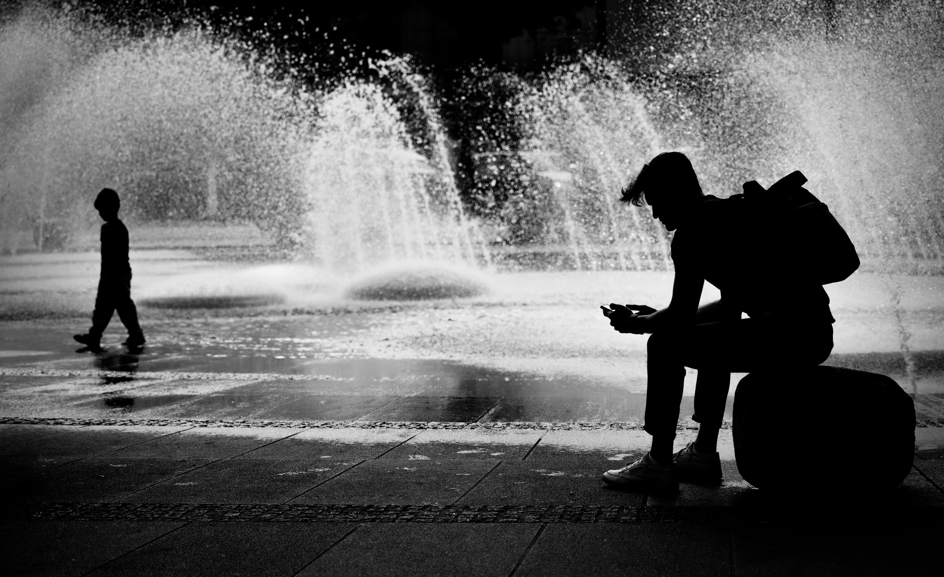 Silhouette sitting by fountain checking phone while child plays in water spray — black and white street photography