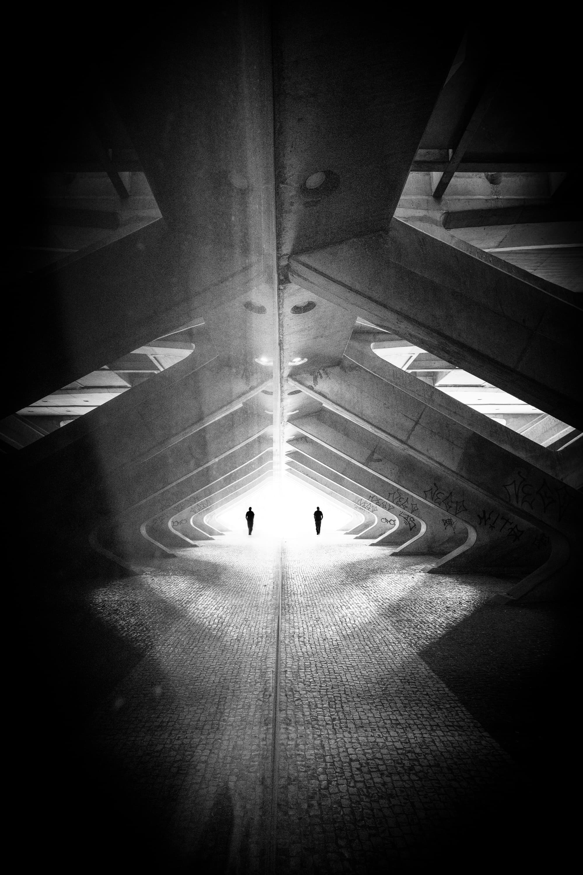 Two silhouettes walking through brutalist concrete underpass with triangular geometry — black and white street photography