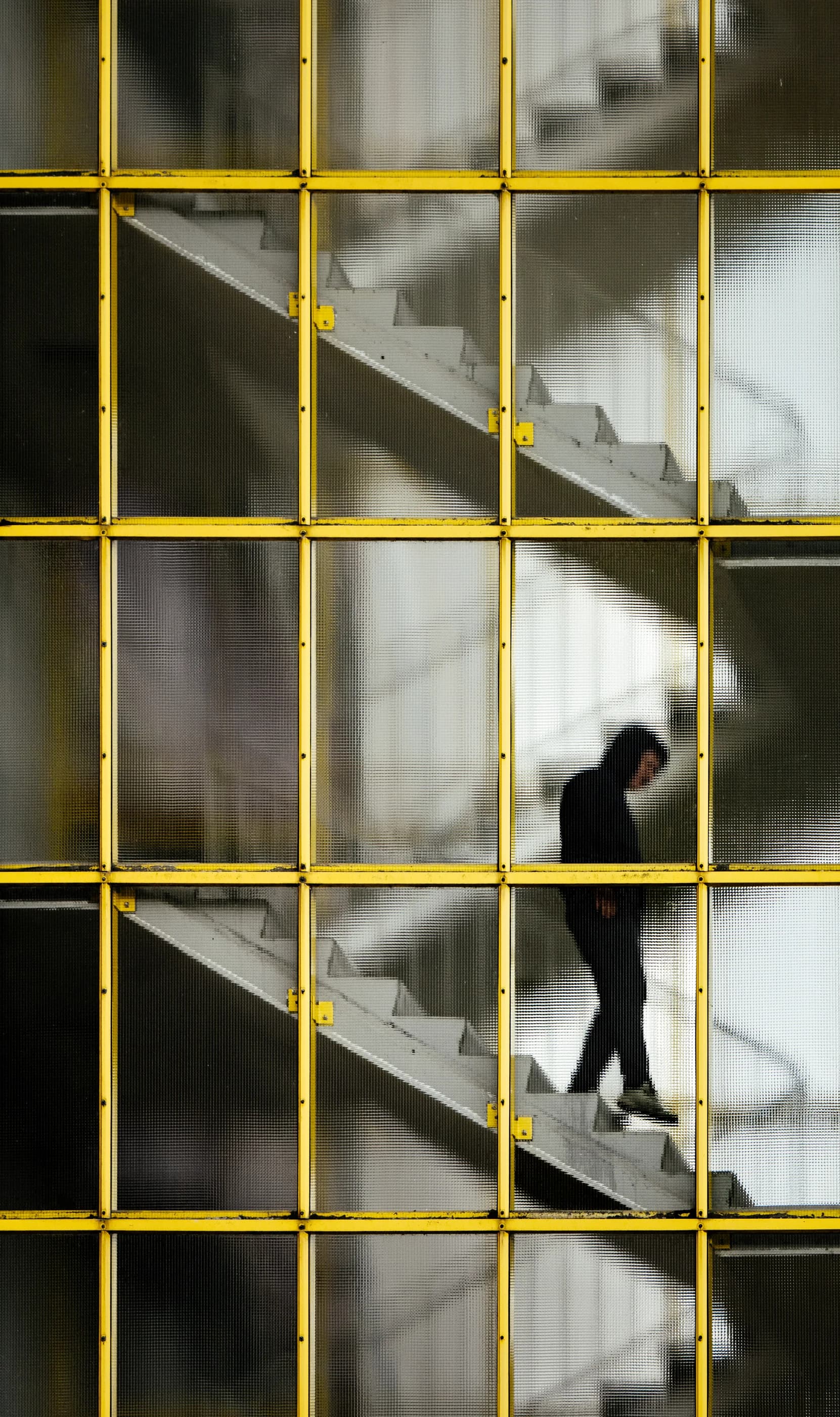 Eine Gestalt mit Kapuze geht eine Treppe hinab, hinter einer Fassade aus gelben Stahlrahmen und Strukturglas im Berliner Hansaviertel